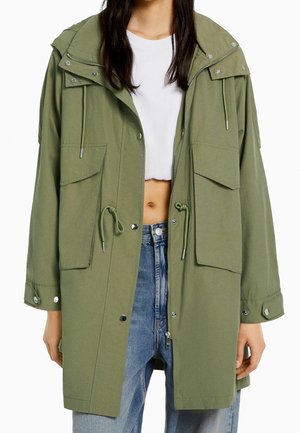 Femme portant une parka à capuche vert olive clair avec de grandes poches avant, un top court blanc et un jean bleu sur un fond uni.