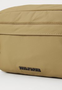 Borsa a spalla in nylon beige con una texture liscia, dotata di una chiusura a zip nera e logo "HILFIGER" impresso sul lato.