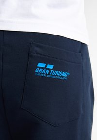 Námořnické modré kalhoty se zadní kapsou s modře potisknutým logem "GRAN TURISMO® THE REAL DRIVING SIMULATOR". Bavlněný materiál.