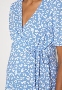 Primo piano di una donna incinta che indossa un vestito avvolgente blu con stampa floreale bianca e un nastro legato in vita.