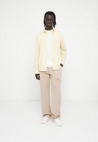 Lichtgeel gestreept overhemd met knopen over een wit T-shirt, gecombineerd met beige wijde broek met steekzakken aan de voorkant en logodetail.