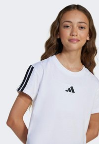 White cotton T-shirt featuring a black embroidered Adidas logo and three black stripes on each shoulder. Short sleeves, crew neckline.