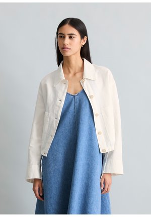 Junge Frau mit langen dunklen Haaren, die eine lockere weiße Jacke mit Knöpfen über einem blauen ärmellosen Jeanskleid trägt und vor einem schlichten grauen Hintergrund steht.