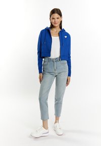 Hoodie blu cropped con cordini e logo a forma di cuore. Indossato sopra una maglietta bianca, abbinato a jeans chiari a vita alta e sneakers bianche.