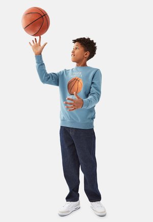 Helles blaues Sweatshirt mit Basketballgrafik und Text, kombiniert mit dunkelblauen Jeans und weißen Sneakers. Basketball auf einem Finger balanciert.