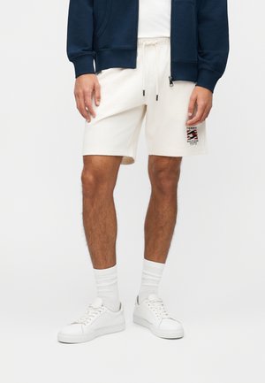 Mężczyzna w kremowych szortach ze sznurkiem i logo Tommy Hilfiger, białych skarpetkach, białych trampkach oraz granatowej kurtce na suwak, na jednolitym tle.