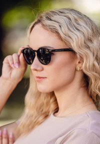 Černé kulaté sluneční brýle s matným povrchem, značka VUCH na nožičkách a odrazové čočky. Vlasy vlnité blond, částečně viditelné.