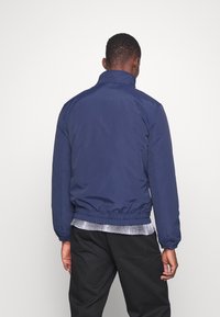 Granatowa lekka kurtka z elastycznymi mankietami i dołem, z kołnierzem typu stójka i gładką fakturą. Noszona na koszuli z kołnierzykiem.