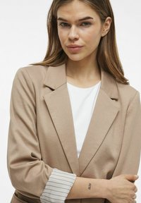 Blazer beige con un design su misura, caratterizzato da una texture liscia e un dettaglio a righe sui polsini. Indossato sopra un top bianco, con una silhouette aderente.