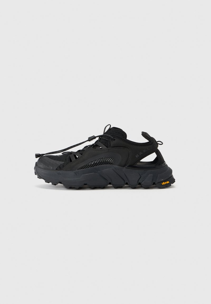 Sapatilhas de caminhada pretas apresentam um cabedal em malha com detalhes em borracha, sola grossa e texturizada, e um sistema de atacadores integrado para um ajuste seguro. Logótipo da Vibram visível.