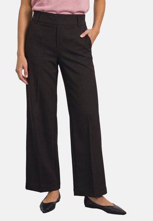 Femme portant un pantalon noir à jambes larges, un haut rose et des ballerines pointues noires, debout avec une main dans la poche sur fond blanc.