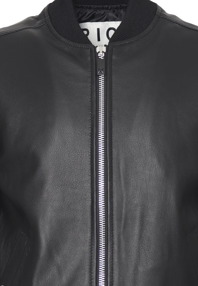 Giubbotto di pelle nera con zip centrale argentata e colletto nero a coste, che mostra parzialmente l'etichetta interna bianca con testo nero.