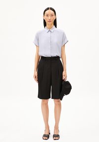 Helles graues Kurzarmhemd, kombiniert mit schwarzen maßgeschneiderten Shorts, die Falten und ein elegantes Design aufweisen. Schwarze Mütze wird in einer Hand gehalten.