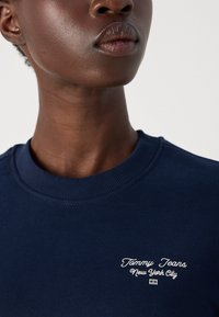 Marinblå hettegenser med ribbestrikket rund hals, med hvit brodert tekst "Tommy Jeans New York City" på venstre brystområde.