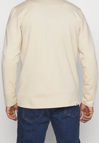 Camicia a maniche lunghe di un leggero color beige, con scollo a girocollo, realizzata in un tessuto morbido. Caratterizzata da una vestibilità comoda e cuciture discrete all'orlo.