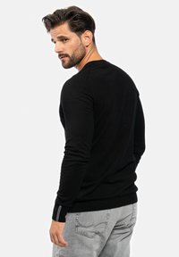 Schwarzer Pullover mit langen Ärmeln aus weichem Material und rundem Halsausschnitt. Auffällige Nähte an den Schultern und Manschetten, kombiniert mit hellgrauer Jeans.