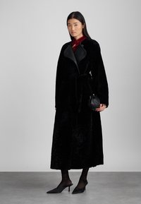 Μαύρο μακρύ παλτό με πλούσια υφή, διαθέτει μαύρο δερμάτινο γιακά και ταιριαστό ζώνη. Το μοντέλο φοράει μαύρες γόβες και κρατάει μια μικρή μαύρη τσάντα.