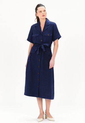 Marineblauwe linnen jurk met korte mouwen, knoopsluiting, twee borstzakken, een strikceintuur en een volle rok. Gekocht met crèmekleurige blokhak schoenen.