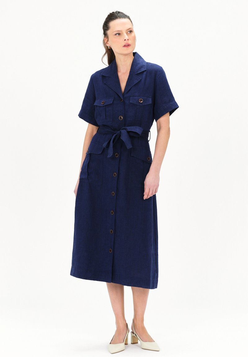 Vestido camisería de lino azul marino con mangas cortas, cierre de botones, dos bolsillos en el pecho, un cinturón de lazo y una falda amplia. Combinado con zapatos de tacón bloque en color crema.