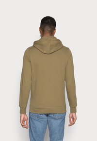 Sudadera con capucha de color verde olivo con una textura suave, puños ajustados y un bolsillo tipo canguro. Se lleva con unos vaqueros de color azul claro.