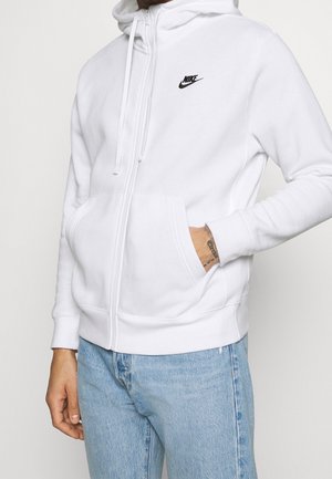 Person trägt weißen Nike Kapuzenpullover mit Reißverschluss und Fronttaschen, eine Hand in der Tasche, kombiniert mit hellblauen Jeans, sichtbares Tattoo am Handgelenk.