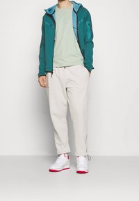 Person som bär en turkos zip-up jacka, ljusgrön t-shirt, vita sweatpants och vita sneakers med röda sulor, stående mot en slät vägg.