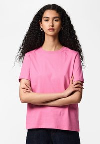 Femme aux longs cheveux bouclés portant des boucles d'oreilles créoles argentées et un t-shirt rose à manches courtes, les bras croisés, debout devant un fond blanc.