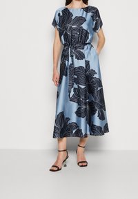 Robe midi florale en satin bleu clair avec un motif de feuilles bleu foncé, manches courtes, taille élastique et détail de ceinture. Talons à brides noirs.