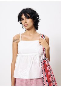 Witte katoenen top met dunne banden, versierd met bloemengen borduursel en een geraffineerde taille. Model houdt een meerkleurige gestreepte totebag vast.