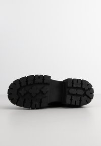 Semelle de chaussure en caoutchouc noir avec des crampons profonds et des motifs de traction en forme de croix et rectangulaires, affichée vue de dessous sur une surface blanche.