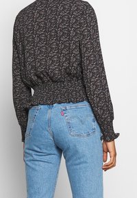 Blusa nera con motivo floreale, con vita arricciata e maniche lunghe, abbinata a jeans azzurri a vita alta con tasche posteriori e dettagli di cucitura.