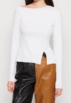Femme portant un haut blanc à manches longues avec un ourlet asymétrique et un pantalon en cuir bicolore noir et marron.