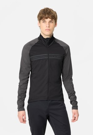 Junger Mann mit einer schwarz-grauen Jacke mit Reißverschluss und langen Ärmeln sowie dunkler Hose, stehend vor einem schlichten weißen Hintergrund.