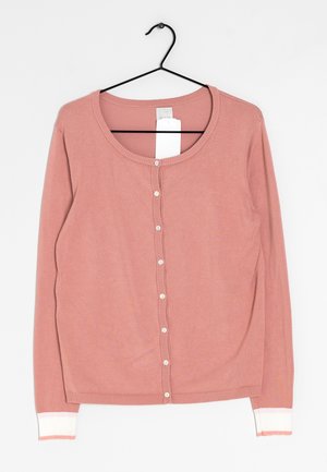 Růžový cardigan s dlouhým rukávem a kulatým výstřihem, na knoflíky, s žebrovanými manžetami a hladkou texturou, zavěšený na černém háčku.