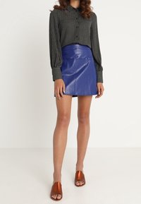 Schwarz-weiße gestreifte Hemdbluse mit langen Ärmeln, kombiniert mit einem blauen Kunstleder-Minirock und braunen Riemchensandalen.