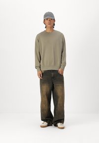 Oversized olijfgroene sweatshirt, donker vervaagde wijde jeans, grijze pet en witte sneakers met beige zolen, staande tegen een neutrale achtergrond.