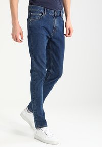 Blå denimjeans med slim fit, med knapp- och dragkedjeflygt, framfickor och subtila sömnadsdetaljer. Bärs med vita sneakers.