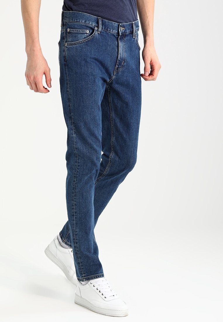 Blå denimjeans med slim fit, med knapp- och dragkedjeflygt, framfickor och subtila sömnadsdetaljer. Bärs med vita sneakers.