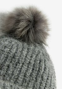 Šedá pletená čepice s měkkým, nadýchaným šedým bambulí na vrcholu. Strukturovaný vzor se skládá z vyvýšených, žebrovaných stehů.