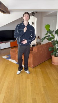 Pantaloni neri, felpa blu navy con cerniera e strisce bianche, scarpe argento. Ambiente interno con divano marrone testurizzato e pianta sullo sfondo.