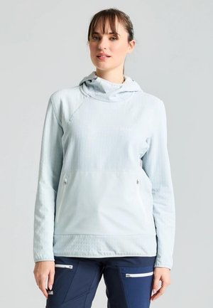Una mujer luce una sudadera con capucha de color azul claro y unos pantalones azul marino con bolsillos con cremallera, sobre un fondo liso y claro.