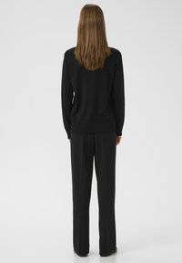 Eine Frau mit glattem braunem Haar steht mit dem Rücken zur Kamera, trägt einen schwarzen Pullover und schwarze weite Hosen vor einem grauen Hintergrund.