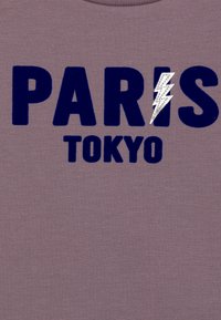 Lilla bomulls T-skjorte med fet marineblå tekst "PARIS" og "TOKYO," med et sølv lynbolt aksent på bokstaven "R."