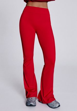 Personne portant un pantalon évasé rouge taille haute et des chaussures de sport argentées, debout devant un fond gris clair uni.