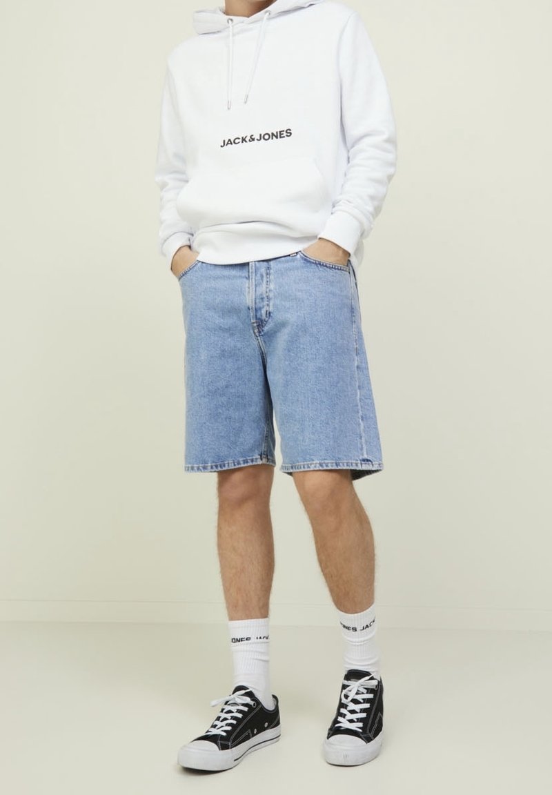 Ljusblå denimshorts, avslappnad passform, med synlig sömnad. I kombination med en vit hoodie med svart logotyp, svarta sneakers och vita strumpor.