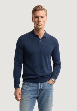 Blonde man die een marineblauw poloshirt met lange mouwen draagt, lichtblauwe spijkerbroek, een zilveren horloge en een armband, staand met een hand in zijn zak.