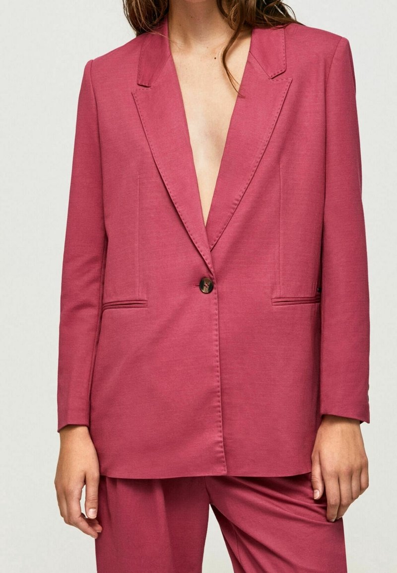 Femme portant un blazer ajusté couleur vieux rose avec un seul bouton et un pantalon assorti, montrée des épaules jusqu'au milieu de la cuisse.