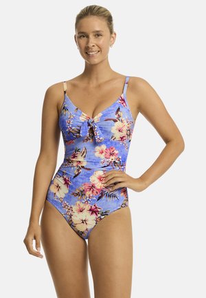 Een vrouw glimlacht terwijl ze een blauwe bloemen-bikini draagt. Haar haar is vastgebonden en ze poseert zelfverzekerd met één hand op haar heup.