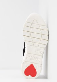 Semelle blanche de basket avec l'inscription "LOVE MOSCHINO" en relief et une forme de cœur rouge près du talon, posée verticalement contre une surface blanche.