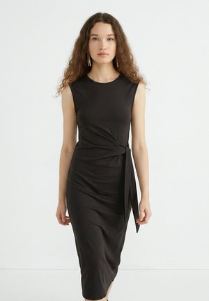 Vrouw met lang krullend haar die een mouwloze zwarte midi-jurk draagt met een knoopdetail aan de zijkant, staand tegen een effen witte achtergrond.
